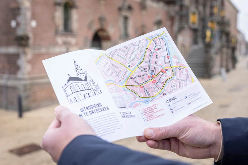 Osoba trzymająca broszurę ze szczegółowym przewodnikiem miejskim z mapą i tekstem napisanym w języku niderlandzkim, stojąca w pobliżu zabytkowego budynku z cegły.