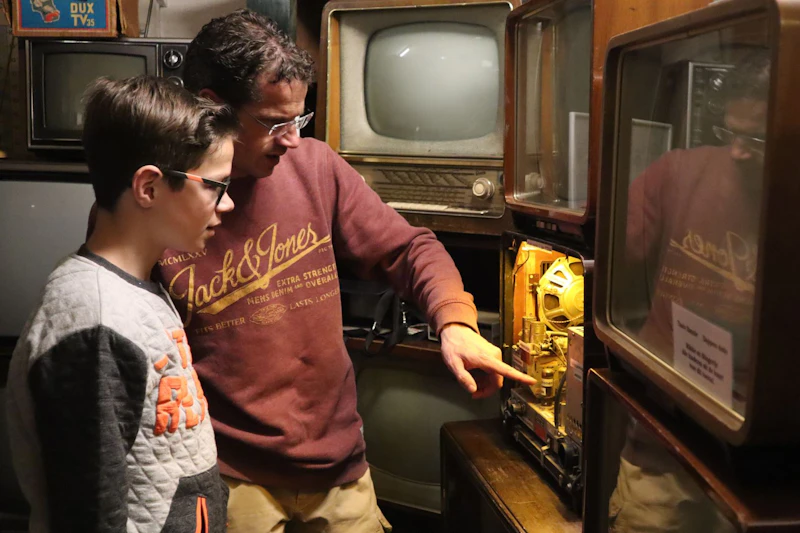 Dorosły i dziecko badają wewnętrzne elementy telewizora vintage pośród innych starych telewizorów w słabo oświetlonym pomieszczeniu.