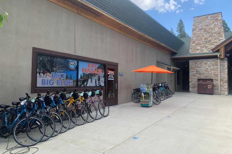 Rowery ustawione przed budynkiem ze znakami „Bike Rentals!” i „Bike Big Blue!” Pomarańczowy parasol osłania więcej rowerów.