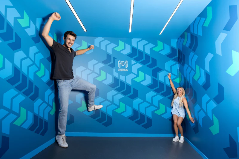 Dwie osoby pozujące w niebieskim pokoju z geometrycznymi wzorami i logo „Muzeum Iluzji” na tylnej ścianie.