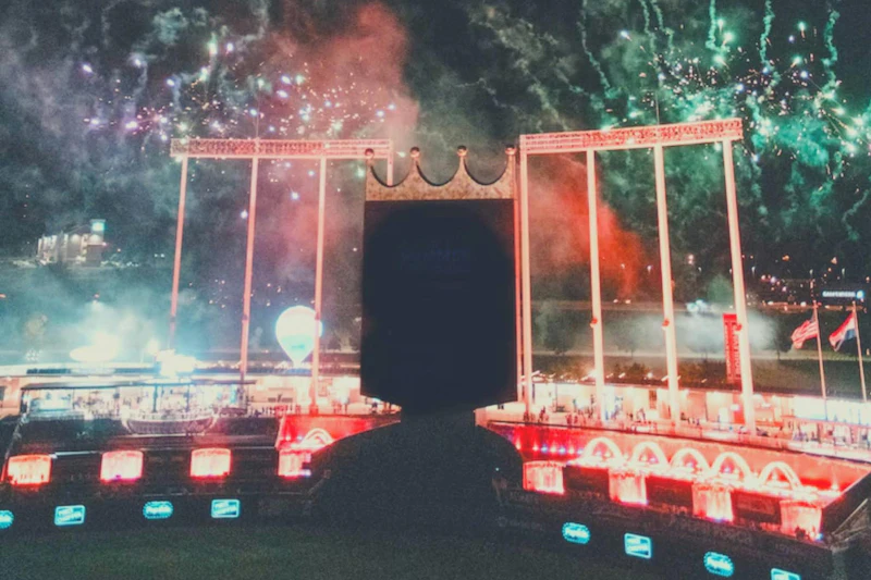 Nocna scena na stadionie z oświetlonymi fontannami, dużym ekranem i fajerwerkami rozświetlającymi niebo nad nami.
