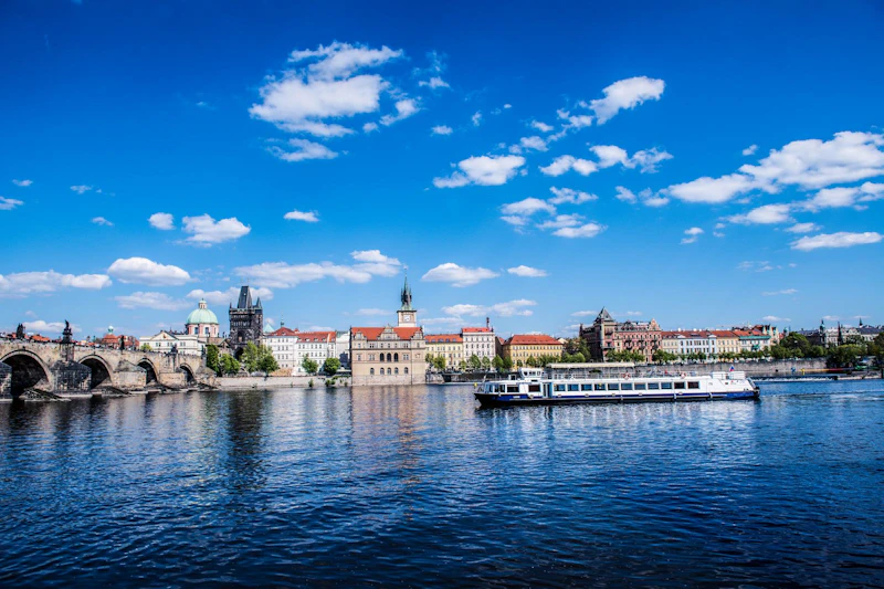 Łódź płynie po rzece z zabytkowymi budynkami, w tym wieżą i kościołem, w tle pod jasnoniebieskim niebem z chmurami.