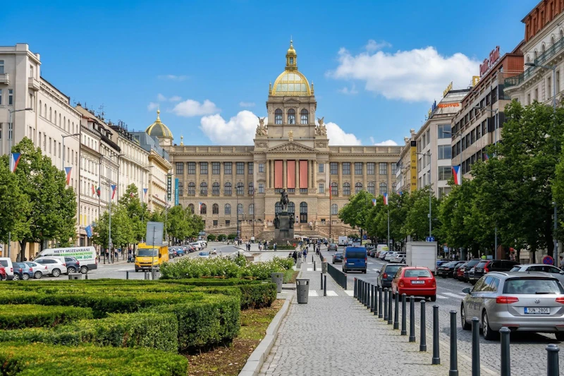 Szeroka ulica z przejściem dla pieszych prowadząca do dużego zabytkowego budynku ze złotą kopułą, w otoczeniu drzew i samochodów.