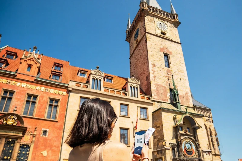 Osoba trzymająca przewodnik audio stoi przed zabytkowym budynkiem z wieżą zegarową pod czystym, błękitnym niebem.