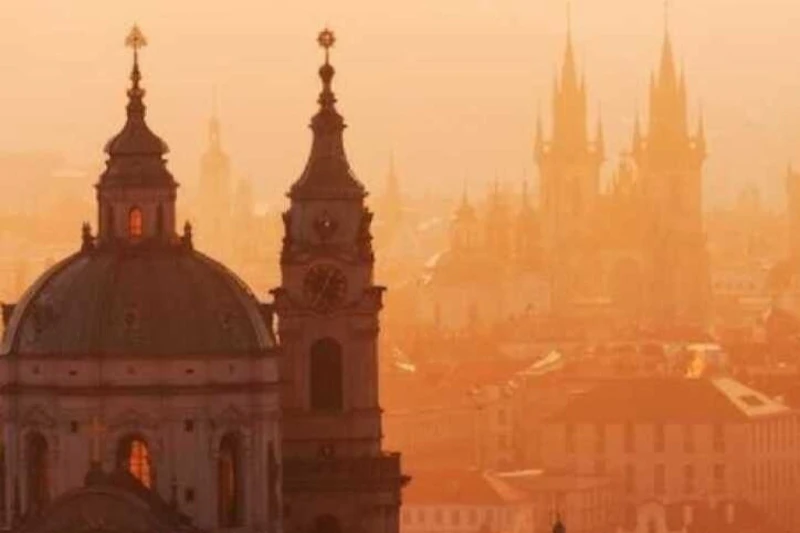 Sylwetki ozdobnych wież i iglic skąpane w ciepłym, pomarańczowym niebie zachodzącego słońca, z budynkami miejskimi w tle.