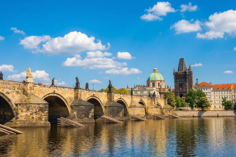 Nad spokojną rzeką rozpościera się zabytkowy kamienny most z posągami, z kopulastymi i iglastymi budynkami pod błękitnym niebem z chmurami.