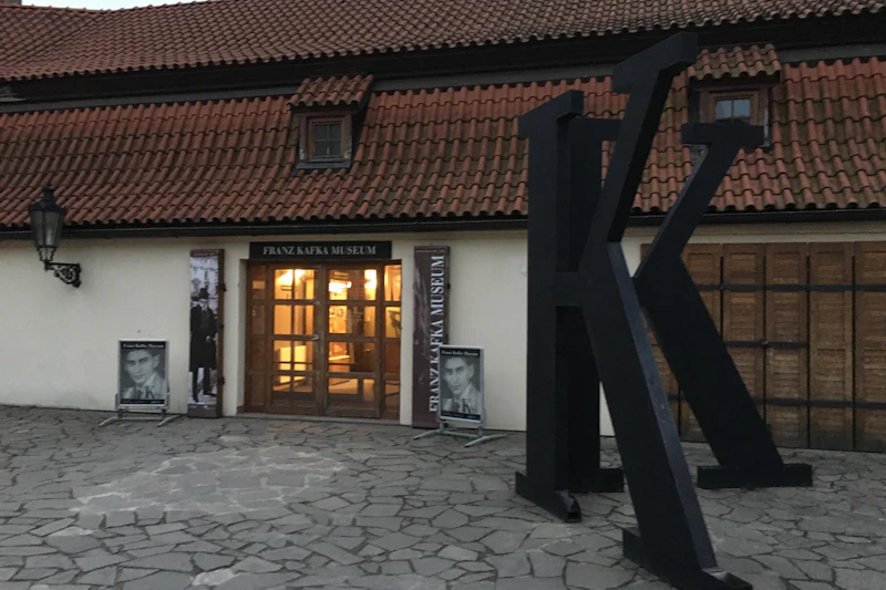 Na zewnątrz Muzeum Franciszka Kafki z dużą czarną rzeźbą „K” z przodu i plakatami przy wejściu pod dach wyłożony dachówką.