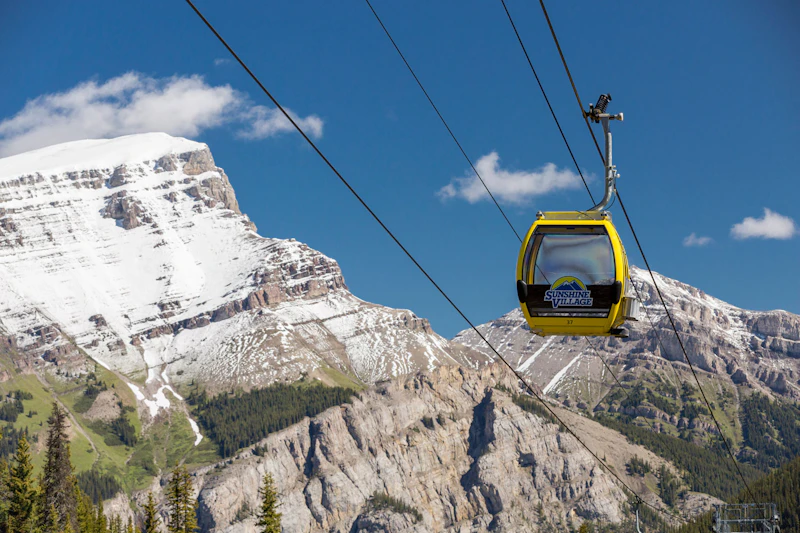 Żółta gondola z napisem „Sunshine Village” jeździ na kablach na tle ośnieżonych gór i błękitnego nieba.