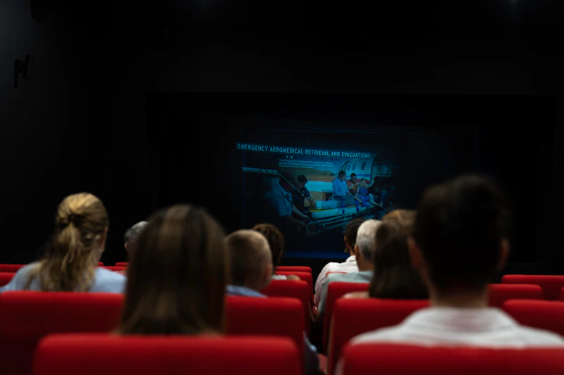 Publiczność siedząca w ciemnym teatrze, oglądająca ekran przedstawiający scenę ewakuacji medycznej.