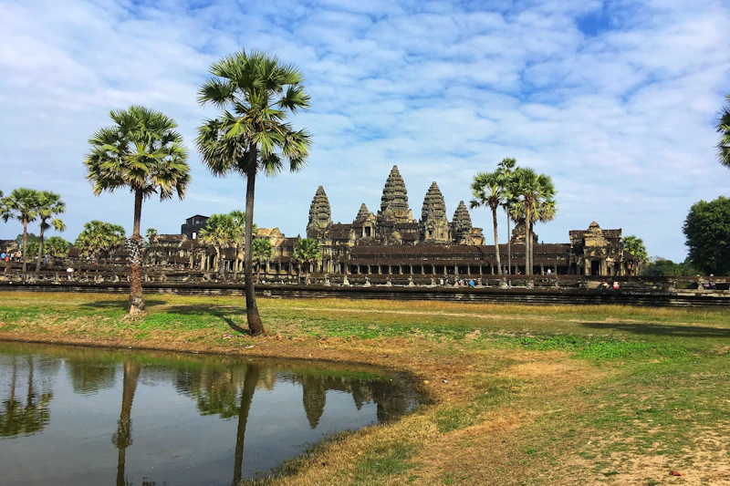 Świątynia Angkor Wat z palmami, trawiastym terenem i lustrzanym stawem pod częściowo zachmurzonym niebem.