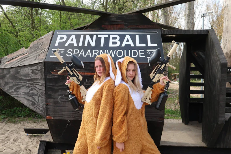 Dwie osoby w brązowych zwierzęcych śpioszkach trzymają pistolety do paintballa przed tabliczką „PAINTBALL SPAARNWOUDE” na zewnątrz.