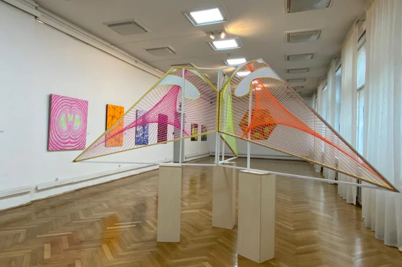 Galeria sztuki z kolorowymi geometrycznymi instalacjami i abstrakcyjnymi obrazami na białych ścianach oraz wypolerowaną drewnianą podłogą.