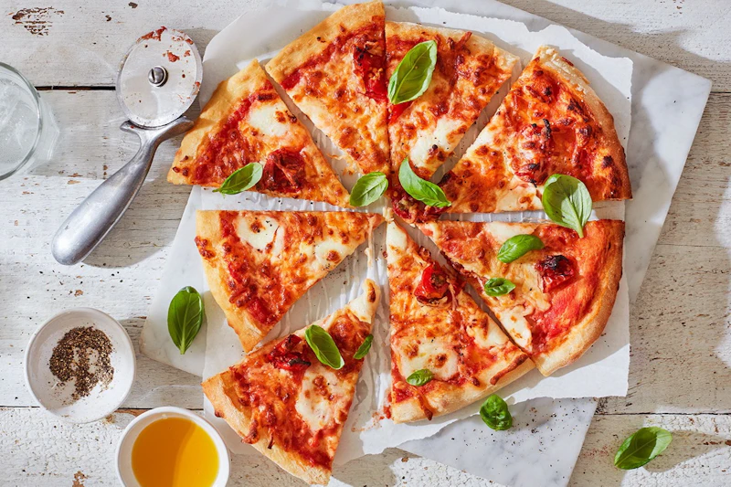 Pokrojona w plastry pizza margherita z liśćmi bazylii, obok foremki do pizzy i małych miseczek z żółtym sosem i oliwą z oliwek na białej powierzchni.