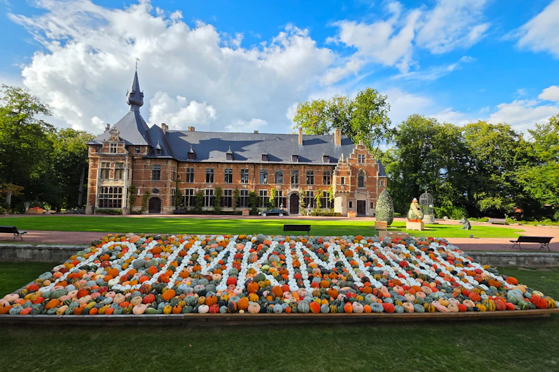 Zabytkowy budynek ze spadzistym dachem za kolorową dynią z napisem „PUMPKINS” pod częściowo zachmurzonym niebem.