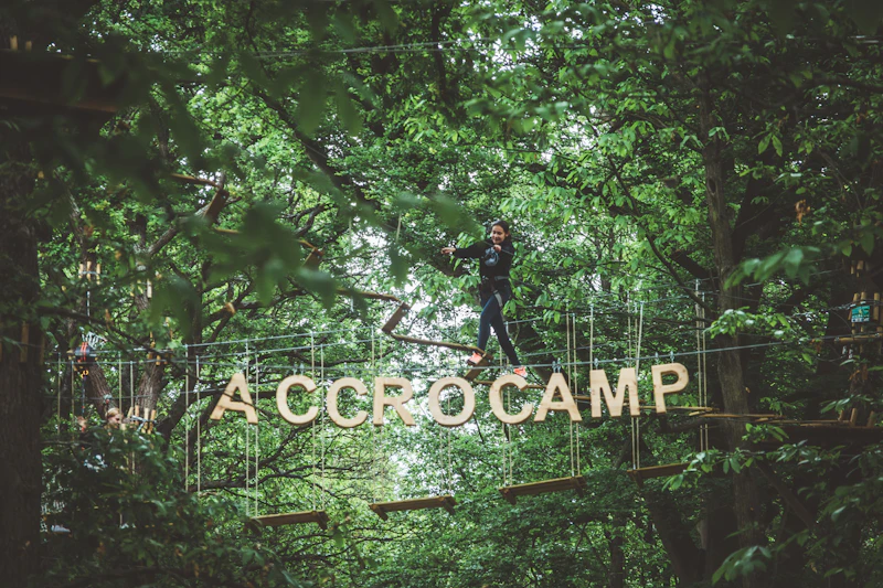 Osoba balansuje na trasie linowej na terenie zalesionym, poniżej wiszą duże litery z napisem „ACCROCAMP”.