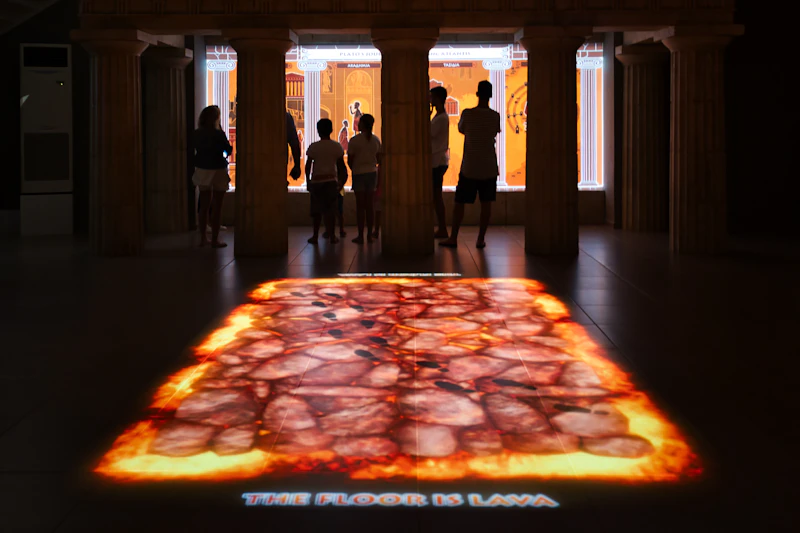 Osoby obserwujące na eksponacie interaktywną ekspozycję przedstawiającą „The Floor is Lava”, z sylwetkami widocznymi na żywym tle.