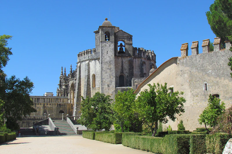 Starożytny kamienny budynek z wieżami i łukami, otoczony drzewami i żywopłotami na zadbanym dziedzińcu pod czystym, błękitnym niebem.