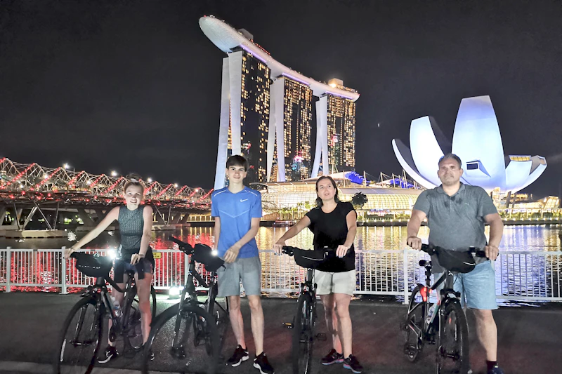 Trzy osoby z rowerami stoją w nocy przy nabrzeżu, z Marina Bay Sands i budynkiem w kształcie lotosu w tle.