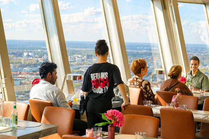 Restauracja z dużymi oknami zapewniającymi widok na miasto. Patroni siedzą przy stołach, a obok przechodzi kelner.
