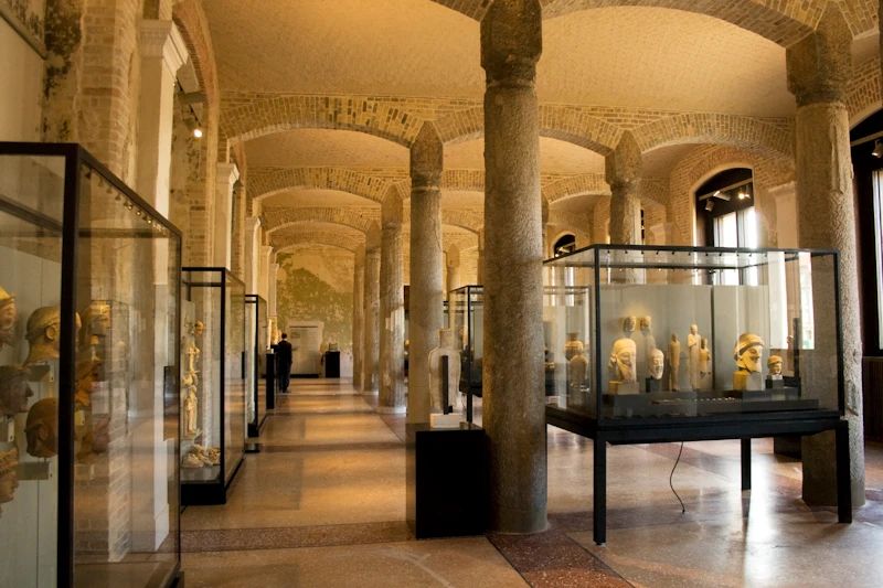 Sala muzealna z kamiennymi kolumnami, łukowymi sufitami i szklanymi gablotami prezentującymi rzeźby i artefakty.