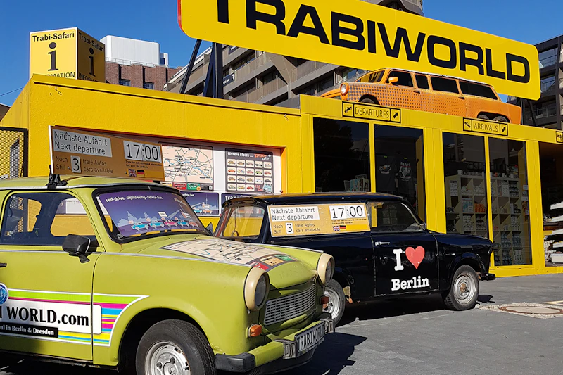 Dwa kolorowe samochody Trabant zaparkowane przed TrabiWorld, żółty budynek z innym Trabantem na dachu pod czystym, błękitnym niebem.