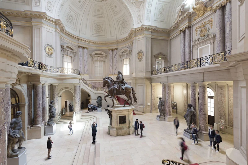 Przestronna sala o klasycznej architekturze, z centralnym pomnikiem jeździeckim i zwiedzającymi obserwującymi okolicę.
