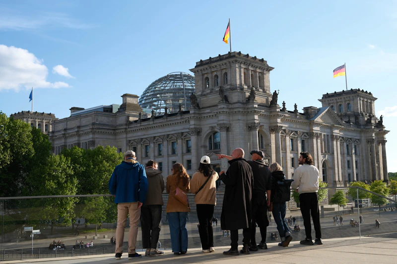 Grupa ludzi stojąca na zewnątrz, zwrócona w stronę budynku Reichstagu ze szklaną kopułą i niemiecką flagą na szczycie.