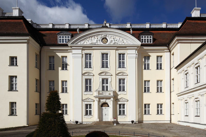 Ozdobny żółty budynek z białymi akcentami, wysokimi oknami i ozdobnym frontonem, pod częściowo zachmurzonym niebem. Małe drzewo na pierwszym planie.