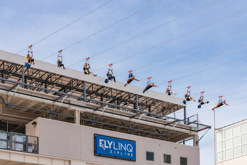 Ludzie zjeżdżają nad budynek ze znakiem „Fly LINQ Zipline” i czystym niebieskim niebem w tle.