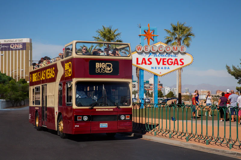 Osoby jadące piętrowym autobusem wycieczkowym przed tablicą „Welcome to Fabulous Las Vegas, Nevada”, z palmami w tle.