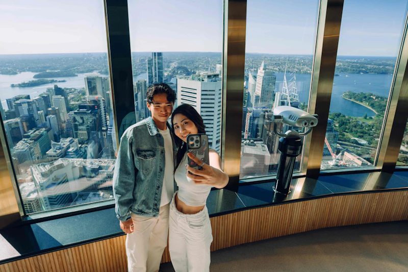 Dwie osoby robią sobie selfie w wieżowcu z dużymi oknami, z widokiem na panoramę miasta z wysokimi budynkami i odległymi widokami na wodę.