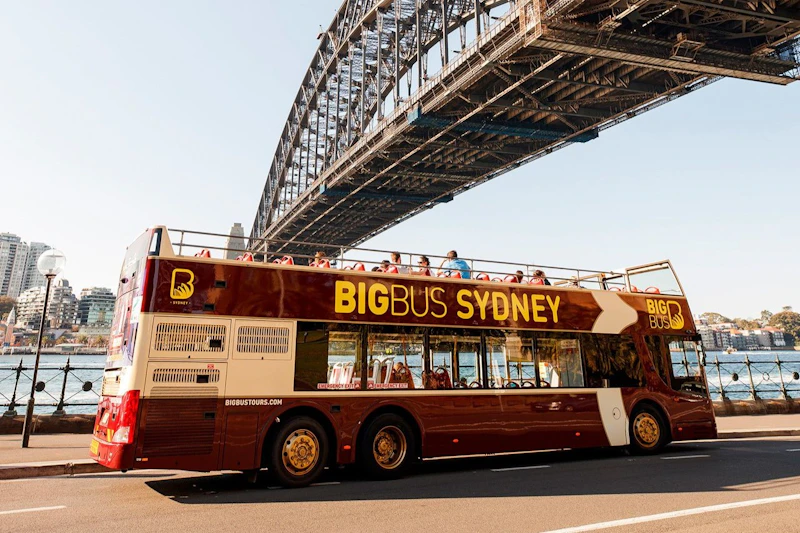 Czerwony piętrowy autobus wycieczkowy o nazwie „Big Bus Sydney” pod mostem portowym w Sydney, z pasażerami siedzącymi na górnym pokładzie.