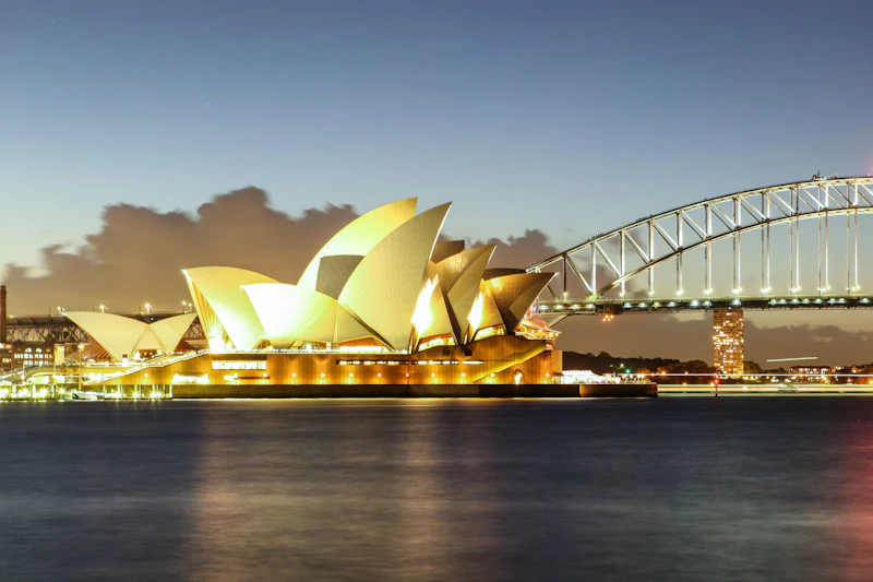 Opera w Sydney oświetlona nocą, z mostem portowym w Sydney w tle i wodą na pierwszym planie.