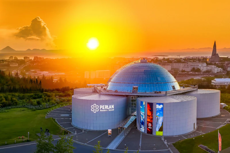 Futurystyczny budynek ze szklaną kopułą oznaczony jako „Perlan Wonders of Iceland” pod żywym pomarańczowym zachodem słońca z pejzażem miasta w tle.