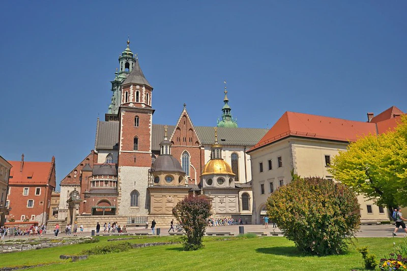 Zabytkowy budynek z wieloma wieżami i kopułami, otoczony trawiastym obszarem z krzewami, na tle czystego błękitnego nieba.