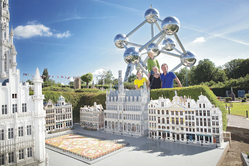 Cztery osoby stojące za miniaturowymi modelami europejskich budynków ze strukturą Atomium w tle w słoneczny dzień.