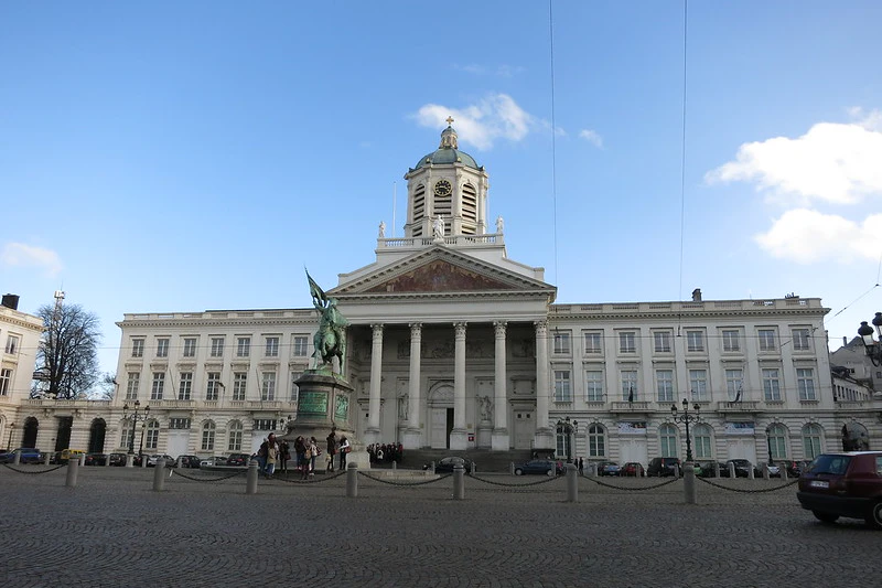 Zabytkowy budynek z kolumnami i kopułą, posągiem z przodu, zgromadzonymi pod nim ludźmi i czystym błękitnym niebem w tle.