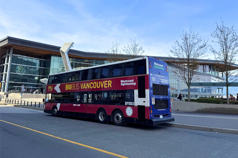 W pogodny dzień przy nowoczesnym budynku stoi piętrowy autobus wycieczkowy o nazwie „Big Bus Vancouver”.