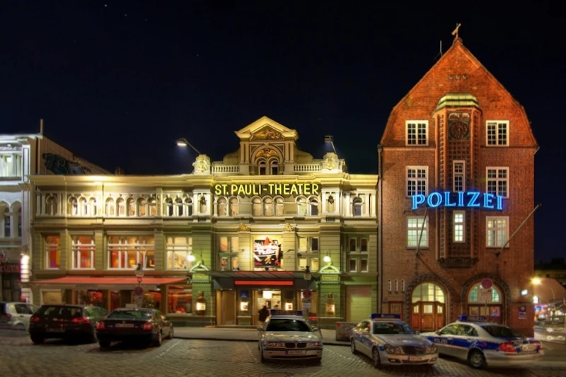 W nocy rozświetlona fasada Teatru św. Pauli znajduje się obok budynku z niebieskim napisem „Polizei”. Przed domem stoi kilka radiowozów.