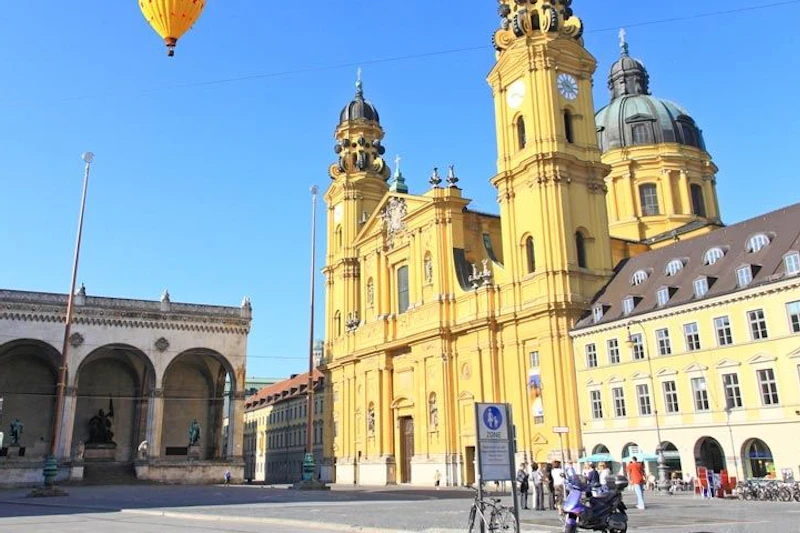Żółty zabytkowy kościół z bliźniaczymi wieżami i kopułą obok budynku z łukowatymi oknami. Na pierwszym planie piesi i zaparkowane rowery.