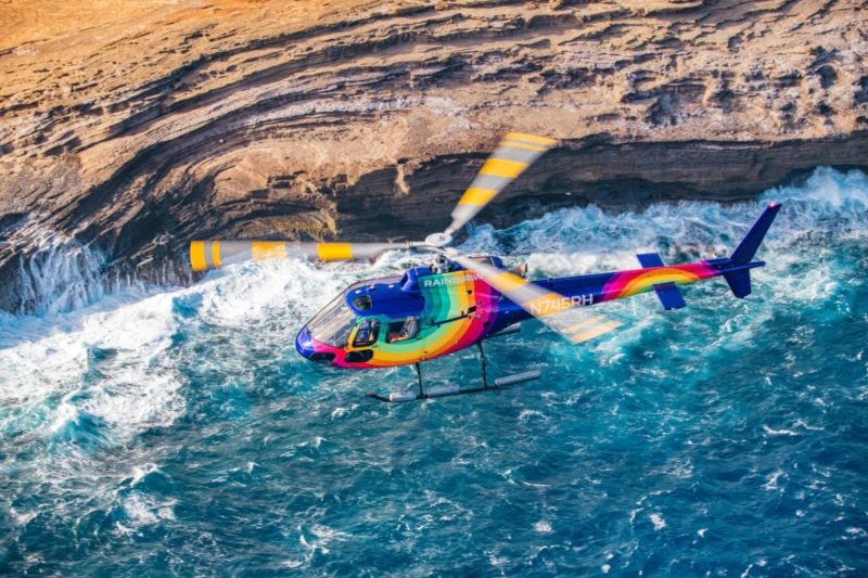 Tęczowy helikopter latający nad oceanem, w pobliżu skalistego wybrzeża klifowego.