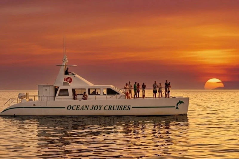 Łódź oznaczona jako „Ocean Joy Cruises” z ludźmi na pokładzie, pływająca po spokojnej wodzie o zachodzie słońca z żywym pomarańczowym niebem.