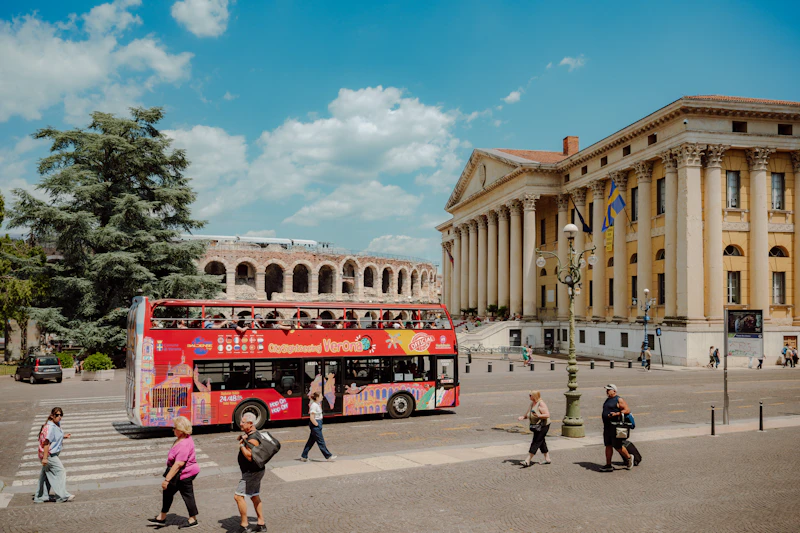 Czerwony piętrowy autobus wycieczkowy przed neoklasycznym budynkiem i starożytnymi ruinami w słoneczny dzień.