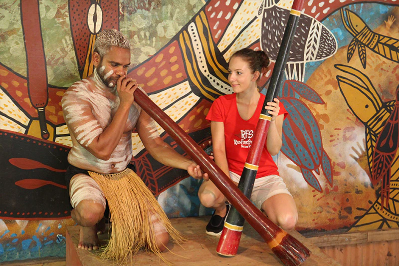 Dwie osoby klękają przed kolorową sztuką aborygeńską. Jeden gra na didgeridoo, podczas gdy drugi trzyma podobny instrument.