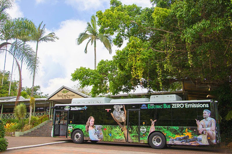 Elektryczny autobus z grafiką dzikiej przyrody zaparkowany przed budynkiem o nazwie „Rainforestation Nature Park” otoczony bujną zielenią.