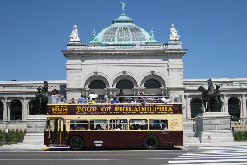 Autobus wycieczkowy z otwartym dachem oznaczony jako „Tour of Philadelphia” przed zabytkowym budynkiem kopułowym z posągami.