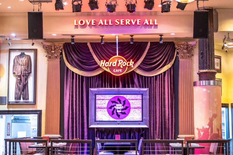 Scena z fioletowymi zasłonami, logo z napisem „Hard Rock Cafe” i frazą „Love All Serve All” powyżej.