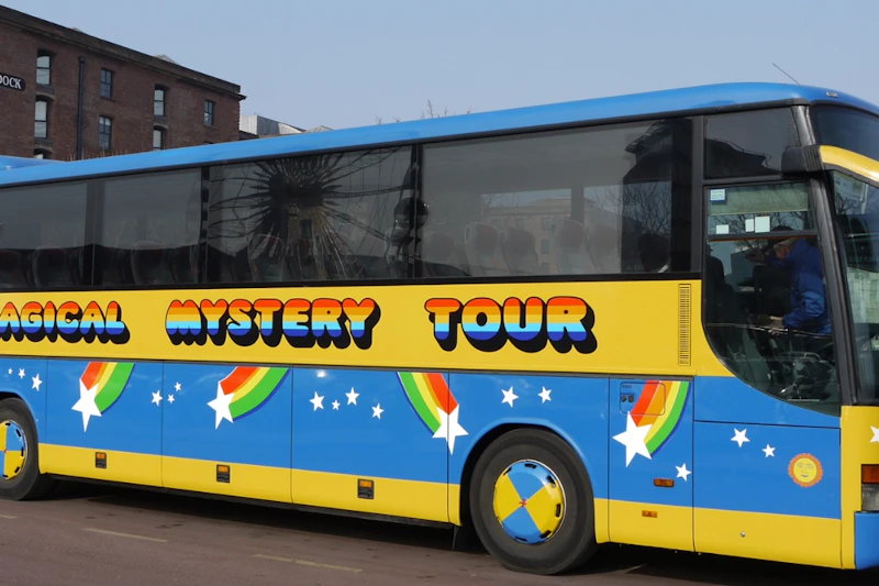 Kolorowy autobus z napisem „Mystery Tour”, ozdobiony tęczami i gwiazdami, zaparkowany na terenie miejskim.