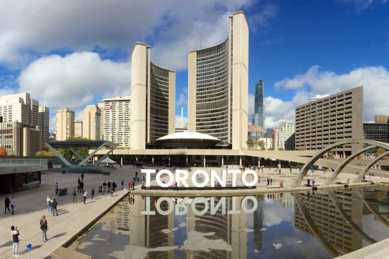 Ludzie spacerują po ratuszu w Toronto z dużym znakiem „TORONTO” i odbijającym się basenem na pierwszym planie.
