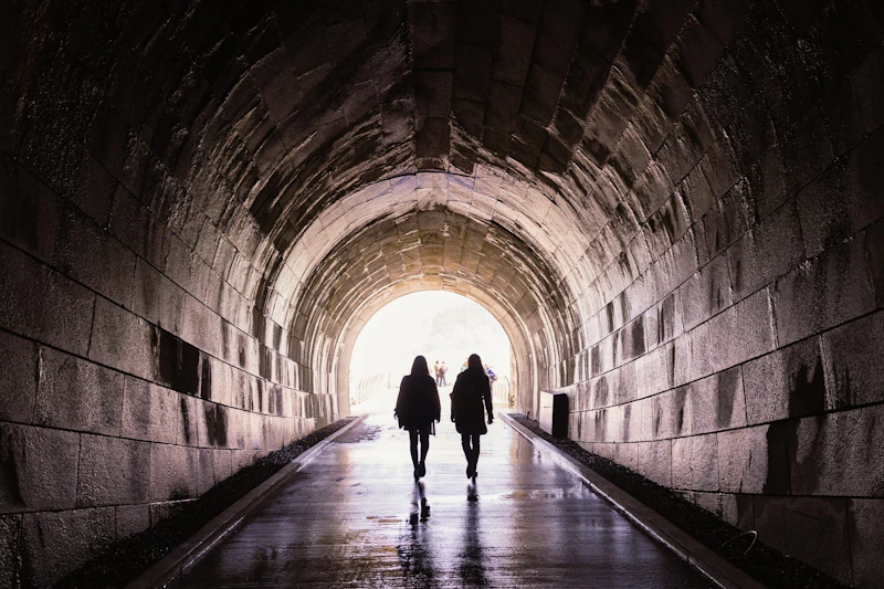 Dwie osoby przechodzą przez kamienny tunel łukowy, kierując się na koniec w stronę jasnego światła. Podłoże jest mokre i odbija światło.
